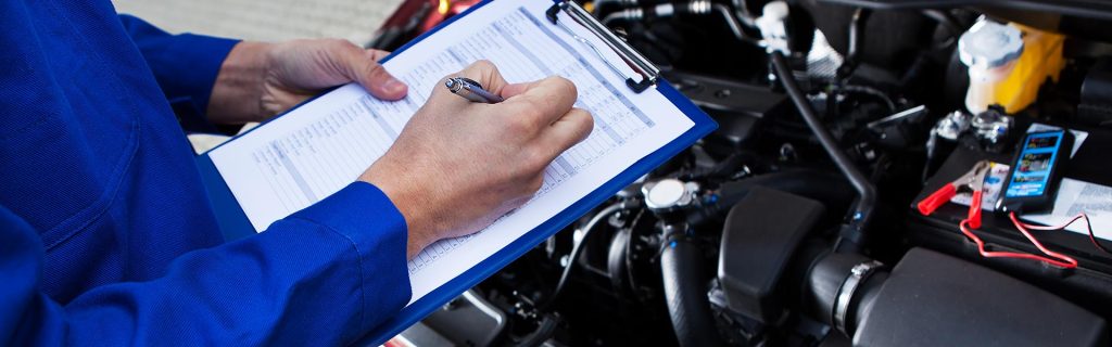 Mechanic in blue coveralls filling out a service checklist over a car engine bay.